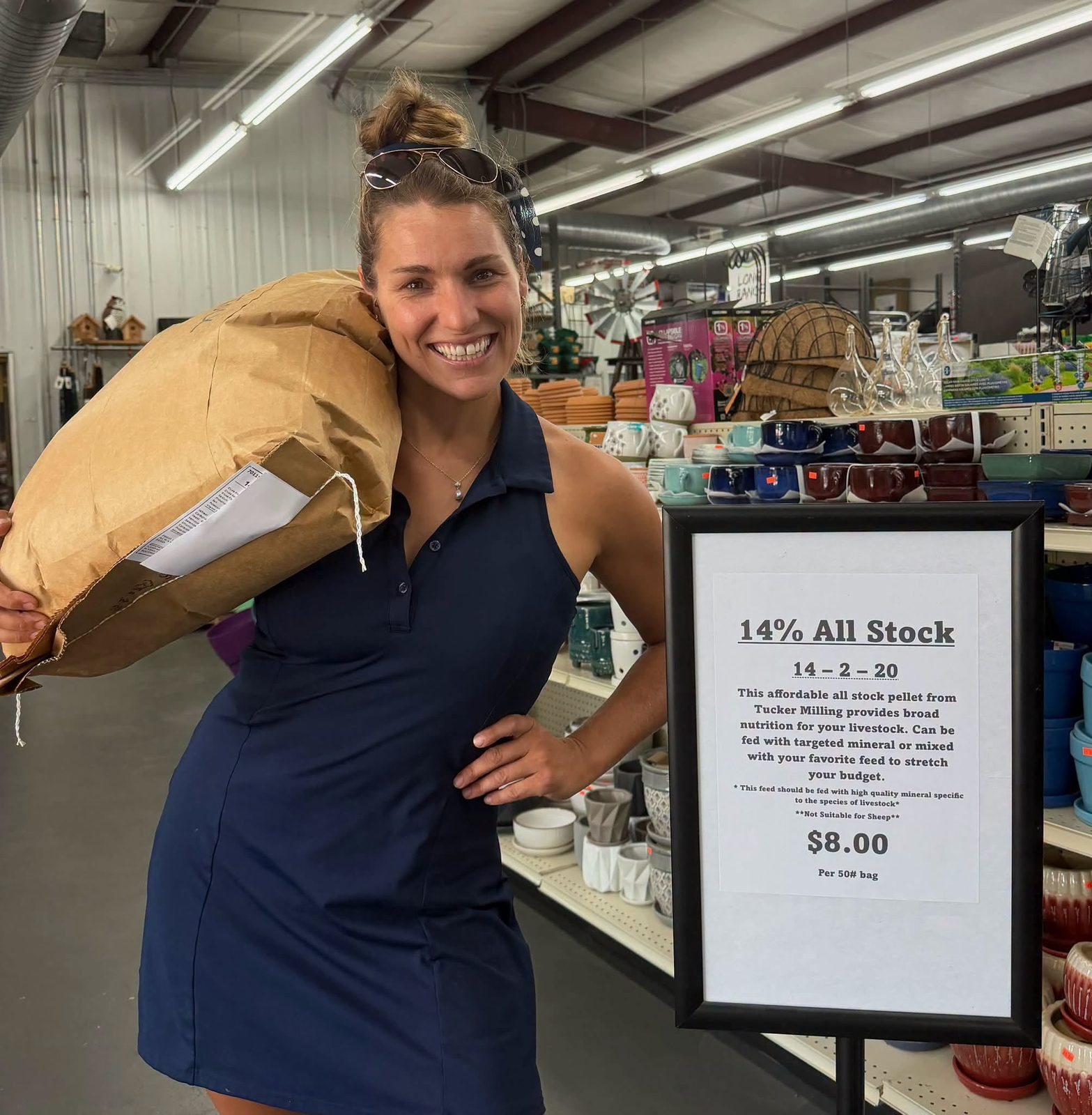 Elisa with a 50 lb Tucker Milling all-stock bag over one shoulder, next to the feed price sign